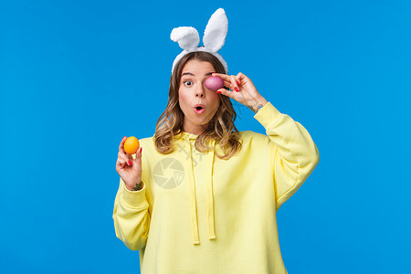 快乐的欧洲金发女子在兔子耳朵里 兴奋和惊讶地拿着两个彩漆的复活节日鸡蛋 蓝背景 Y I L B 教育 有趣的美丽高清图片素材