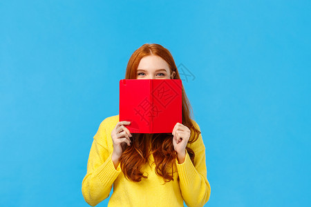 时髦而富有创造力的快乐魅力红发女人 青少年把脸藏在红色笔记本后面 微笑着 狡猾地咧着嘴笑 向朋友隐瞒她在日记中写的东西 站在蓝色背景图片