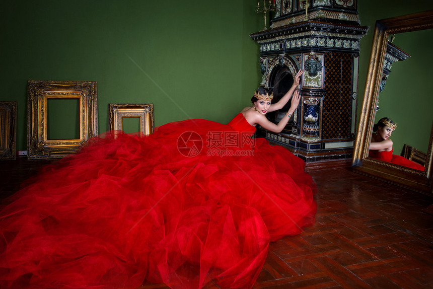 穿长红色裙子的漂亮女人穿着皇家冠在摄影棚豪华室内壁图片
