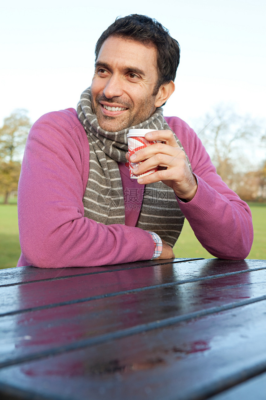 坐在公园桌子旁拿着热饮料的男人图片
