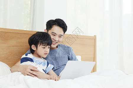 动画片机器猫年轻爸爸陪儿子在床上使用笔记本电脑背景