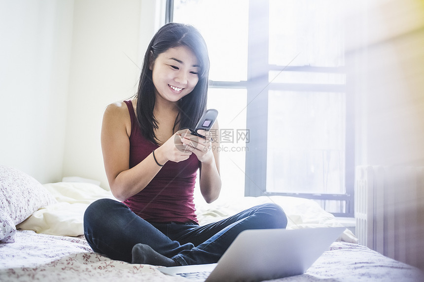坐在床上使用智能手机和笔记本电脑的年轻女性图片