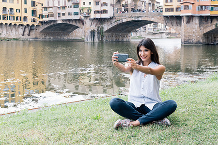坐在河前面的岸上年轻女子使用智能手机自拍图片