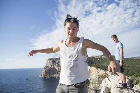 意大利撒丁岛卡波卡西亚一名身穿背心双臂张开站在海边悬崖上跳跃的年轻男子自由高清图片素材