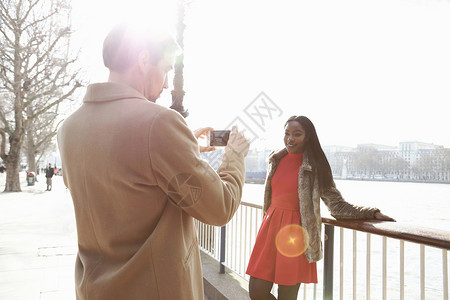 男人给站在河边的年轻女人靠着栏杆拍照片时髦休闲装高清图片素材