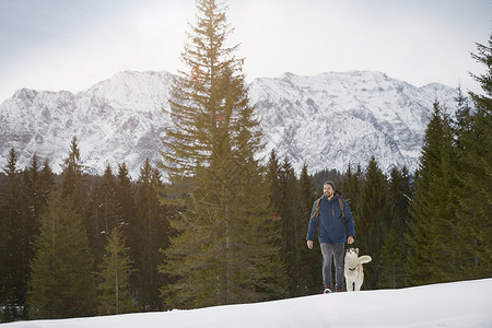年轻人和宠物狗在雪覆盖的山脉上漫步图片