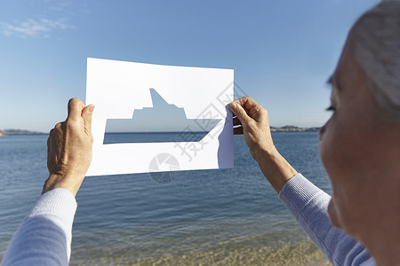站在海对面的老年女子拿着剪出船型洞的纸图片