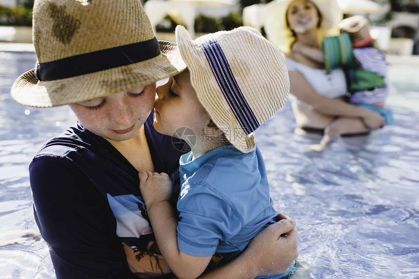 家庭在户外游泳池年轻男孩抱着弟图片