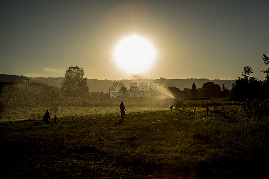 日落时成人和儿童玩农业喷洒器的田地景观图片