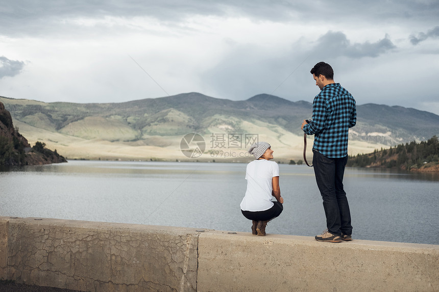 在湖边墙上的男人拿着相机准备给蹲下的女友拍照银角美国科罗拉多州图片