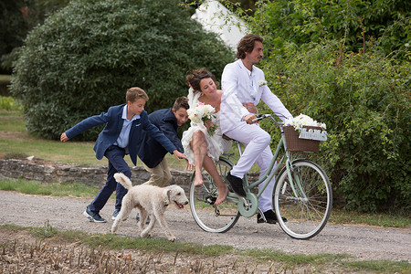 狗兜风年轻的婚礼客人追赶骑自行车的新婚夫妇背景