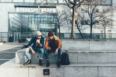 坐在台阶上看着智能手机的两位年轻时尚潮男背景图片