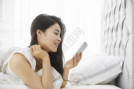 床上拿着手机微笑的女青年枕头高清图片素材