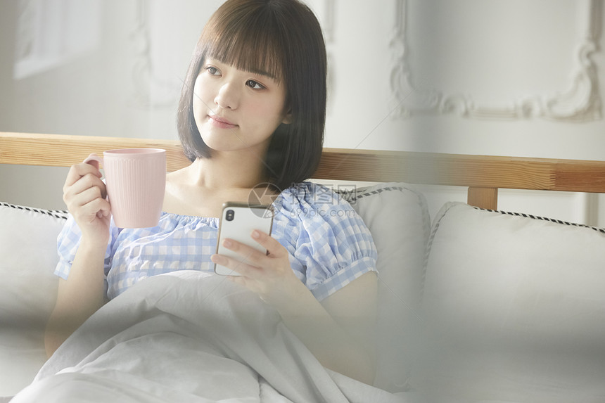 床上拿着水杯和手机的年轻女子图片