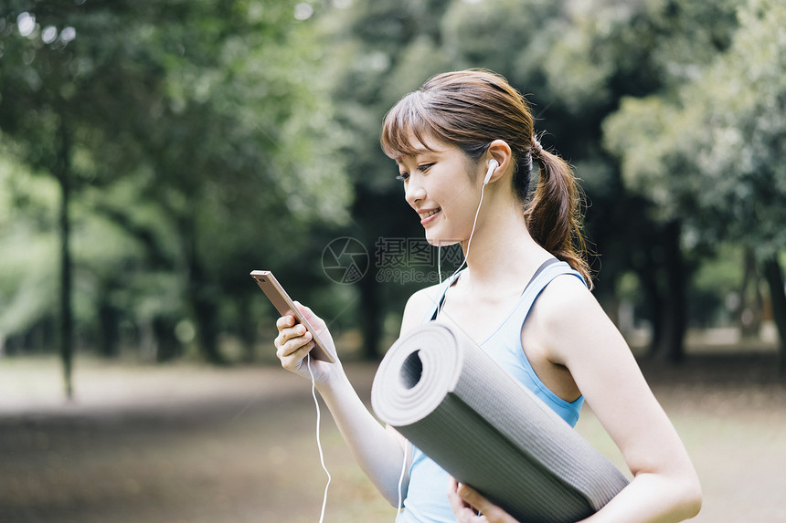 户外抱着瑜伽垫看手机的青年女子图片