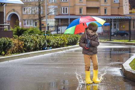 穿着黄靴子的小男孩带着彩虹伞在下雨的公园玩耍图片