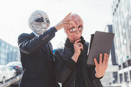 两名戴着外星人面具的男子在城市背光户外使用平板电脑图片