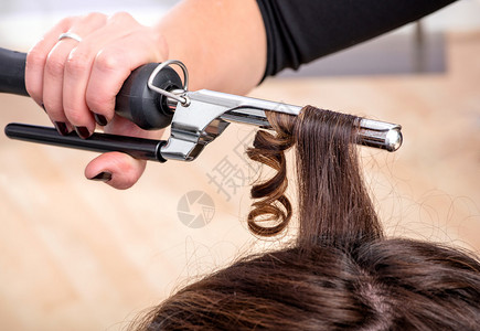发型师在美发沙龙用卷发器或钳子固定女客户棕色长发上的卷发图片