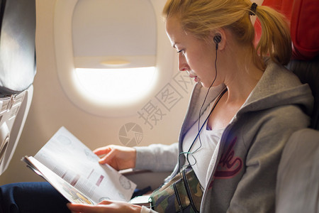 女人在飞机上看杂志和听音乐坐在客舱里看书的女旅行者阳光透过女孩高清图片素材