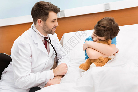 微笑的儿科医生看着小男孩在医院床图片