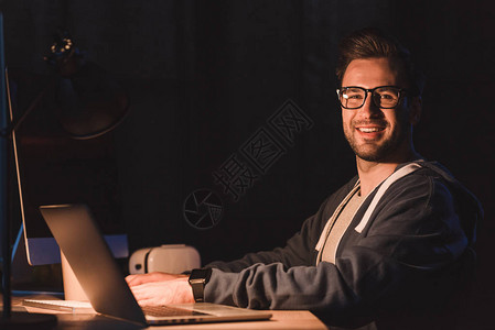 戴眼镜的英俊年轻程序员在夜间使用笔记本电脑和台式计算机时图片