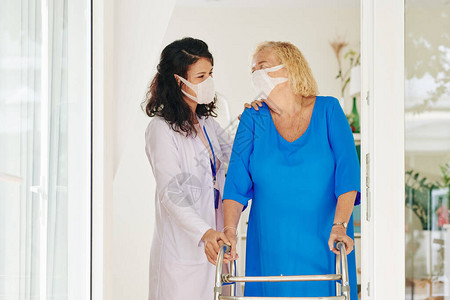 戴医用口罩的护理人员帮助老年妇女在助行器图片