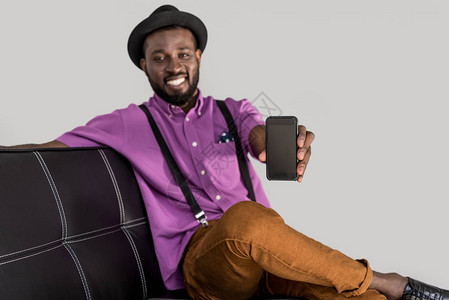 有选择焦点微笑的非洲美国时尚男子显示智能手机在灰色隔离的黑沙发背景图片