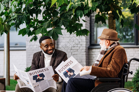 坐在轮椅上幸福快乐的老年残疾人和非洲裔美国人在街上一起阅读报背景图片