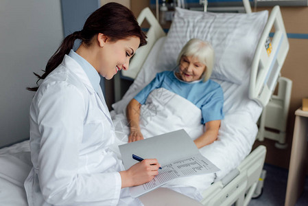 坐在床上并撰写诊断书和在医院睡觉的年长妇女时使用微笑着图片