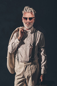 一个有墨镜的时尚胡子老人带着外套和手提箱在反黑背景摄影机上微笑着背景图片