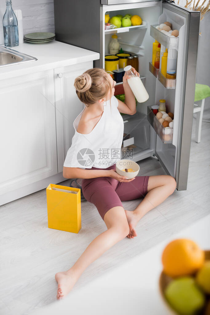 金发年轻女子坐在开着的冰箱附近的地板上时图片