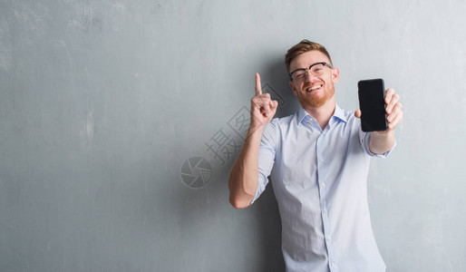 灰色格朗基墙上的年轻红发男子展示智能手机的空白屏幕图片