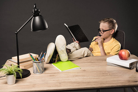 身戴眼镜的男学生用书植物灯彩铅笔苹果钟表和灰底教科书图片