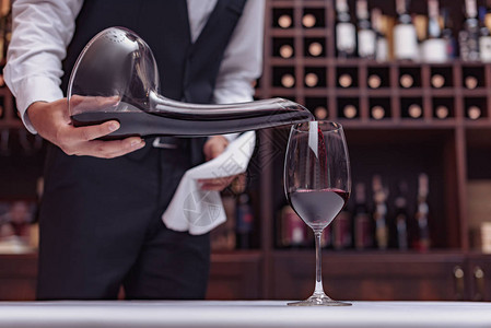 裁剪视图侍酒师将红酒从醒酒器倒入酒窖餐桌上的玻璃杯中背景图片