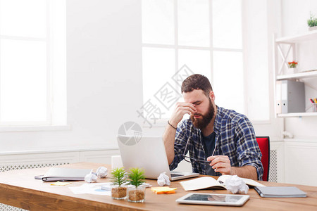 脑阔疼眼睛疼疲惫过度劳累的商人在现代白色办公室内部使用笔记本电脑英俊的男人在随便用电脑工作背景