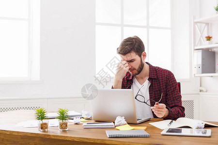 脑阔疼眼睛疼疲惫过度劳累的商人在现代白色办公室内部使用笔记本电脑英俊的男人在随便用电脑工作背景