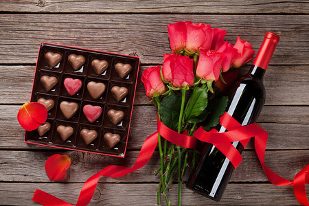 配有红玫瑰和心形巧克力的贺卡以及木本底酒瓶子的红背景图片