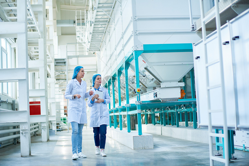 两名身穿实验室大衣的年轻女工在清洁生产车间复制空间中通过电力单位横跨大厅行走的完整图片