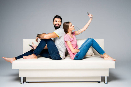 年轻夫妇背靠坐在白沙发上使用在灰色上隔图片