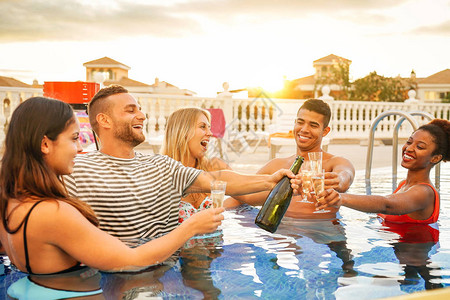 一群快乐的朋友在日落时用一瓶香槟在泳池派对上敬酒年轻人在豪华热带度假村喝起泡酒玩得开心青年背景图片