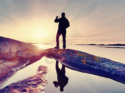 旅游家伙在手机数码相机上拍摄令人惊叹的海景观远足者停留在海平面附近岩石上的镜像池上方一个美丽的日落光创造背背景图片