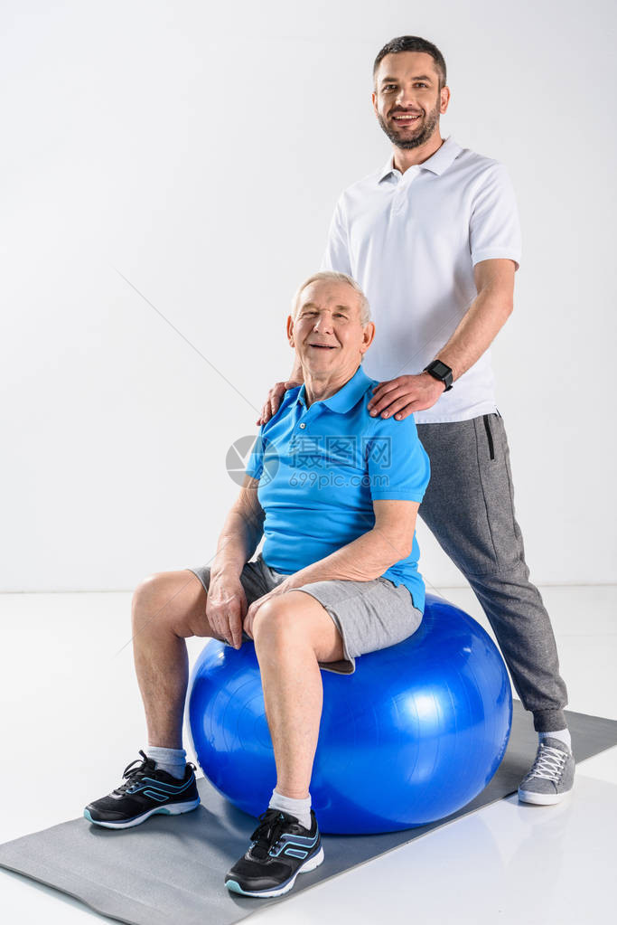 微笑的康复治疗师和健身球上高年级男子在灰色背景图片