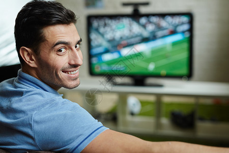 英俊男人微笑的肖像在家里看足球比赛时转图片