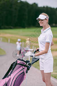 高尔夫球场上戴着帽子和带高尔夫球装备的白色马球微笑的女高尔夫球手图片