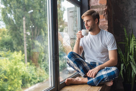 坐在窗台上喝咖啡和在家清晨喝咖啡的年轻男图片