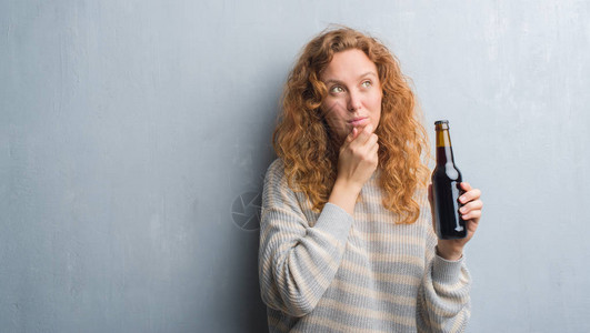 红发女青年在灰色的黑墙上拿着啤酒瓶面对严肃的面孔思考问题图片