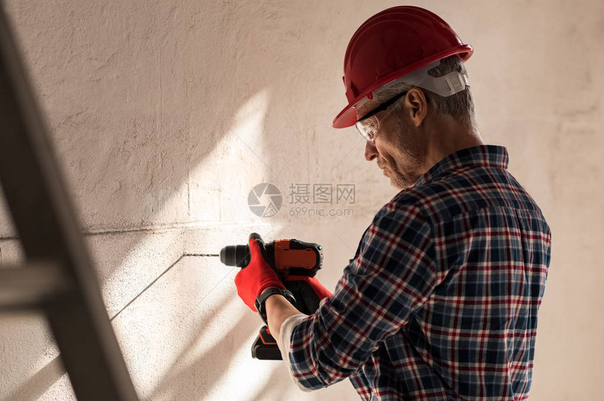 建筑工人用电钻在墙上打洞瓦工在建筑工地的墙上钻孔戴安全帽的成熟男人在墙上打洞图片