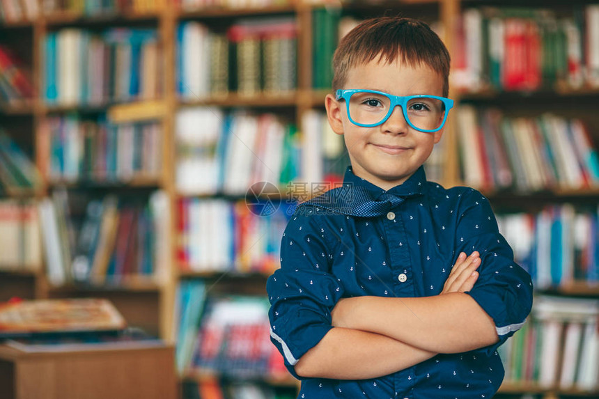 带着蓝色眼镜框和蓝色领带的微笑学生在书本背景的肩膀上男孩在图书馆的多色书架上双手交叉教育图片