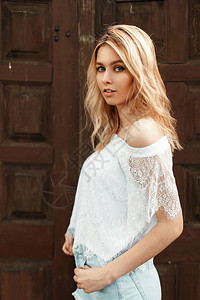 美丽的美国时尚金发美女穿着时髦的白色花蕾上衣在木墙旁背景图片