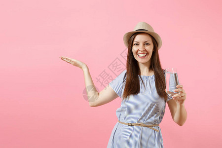 穿着蓝裙子戴帽子和从粉红色背景隔绝的玻璃杯中喝清净纯水的年轻美女肖像图片